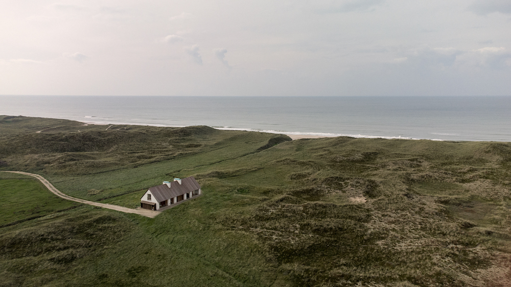Aerial view of a modern guesthouse surrounded by dunes near the sea.